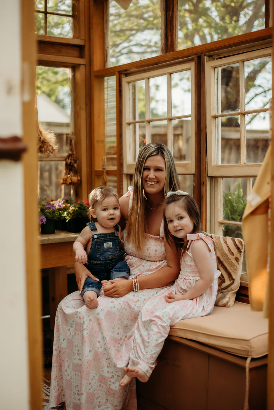Kearstin Zumwalt seated on a window bench inside a greenhouse with her two children.